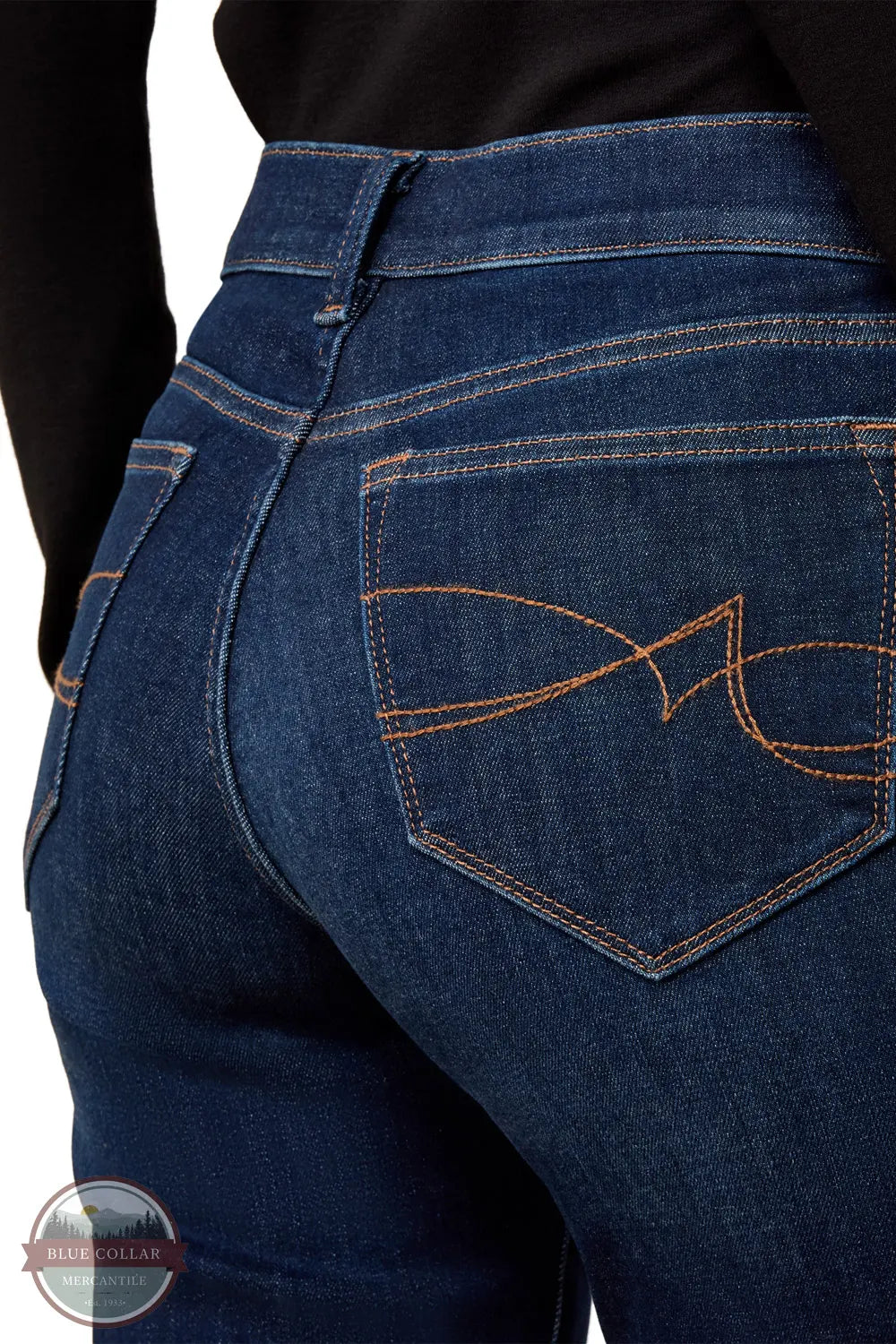 Ariat 10079195 Women's Perfect Rise Vilma Ultrastretch Trouser Jeans in Hayward, Close-up of the back waist and rear pocket of dark blue denim jeans with contrast orange stitching and a butterfly logo on the pocket, worn with a black top.