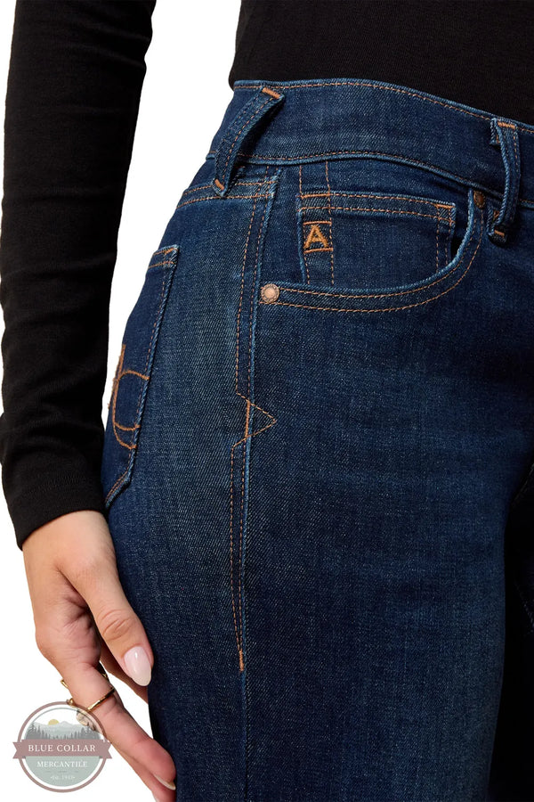 Ariat 10079195 Women's Perfect Rise Vilma Ultrastretch Trouser Jeans in Hayward, Close-up of a person wearing dark blue denim jeans with contrast orange stitching and a small embroidered 'A' logo on the back pocket, paired with a black long-sleeve top against a white background.