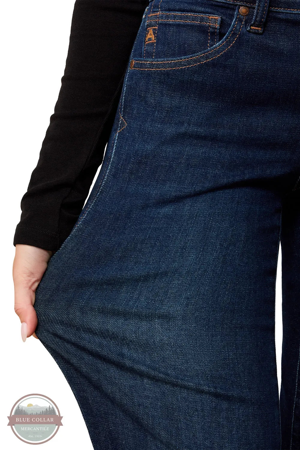 Ariat 10079195 Women's Perfect Rise Vilma Ultrastretch Trouser Jeans in Hayward, Close-up of a person wearing dark blue denim jeans, pulling the fabric outward with one hand to show stretch. A black long-sleeve top is visible, and a small orange 'A' logo appears on the pocket.