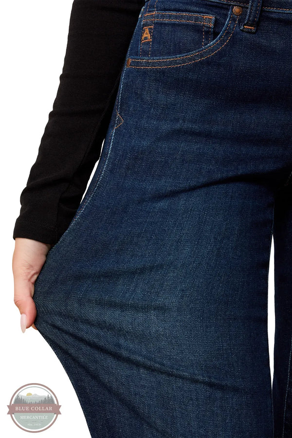 Ariat 10079195 Women's Perfect Rise Vilma Ultrastretch Trouser Jeans in Hayward, Close-up of a person wearing dark blue denim jeans, pulling the fabric outward with one hand to show stretch. A black long-sleeve top is visible, and a small orange 'A' logo appears on the pocket.