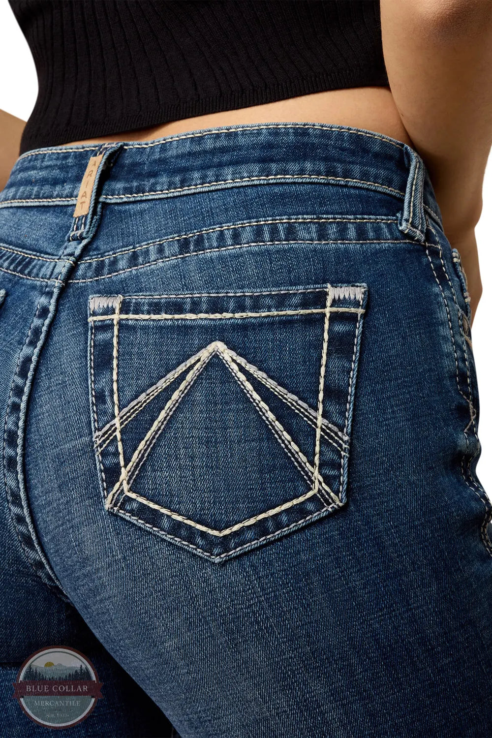 Ariat 10079240 Women's High Rise Teresa Slim Trouser Jeans in Decatur, Close-up of the back pocket of blue denim jeans worn by a model, showing contrast white stitching forming a geometric diamond pattern on the pocket. A black ribbed top is partially visible at the top of the frame.