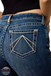 Ariat 10079240 Women's High Rise Teresa Slim Trouser Jeans in Decatur, Close-up of the back pocket of blue denim jeans worn by a model, showing contrast white stitching forming a geometric diamond pattern on the pocket. A black ribbed top is partially visible at the top of the frame.