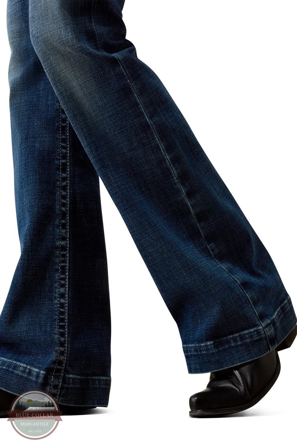 Ariat 10079240 Women's High Rise Teresa Slim Trouser Jeans in Decatur, Close-up of a person's legs wearing dark blue flared jeans and black shoes against a white background.