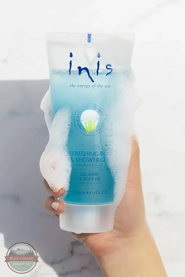 Inis 8005090 Refreshing Bath &amp; Shower Gel 7FL OZ. A hand holding a tube of Inis Refreshing Bath & Shower Gel against a white marble background, with white foam covering the tube.