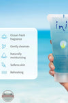 Inis 8005090 Refreshing Bath &amp; Shower Gel 7FL OZ. A hand holds a tube of ini Refreshing Body & Shower Gel against a tropical beach background. A white text box on the left lists product benefits including ocean-fresh fragrance, gentle cleansing, moisturizing, softening skin, and refreshing.
