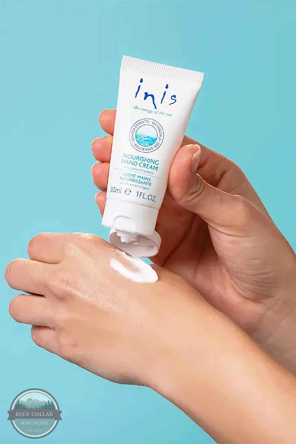 Inis 8015556 Nourishing Hand Cream 2.6FL. OZ. A hand holding a white tube of inis nourishing hand cream over another hand, with white cream being squeezed onto the palm, set against a solid light blue background.
