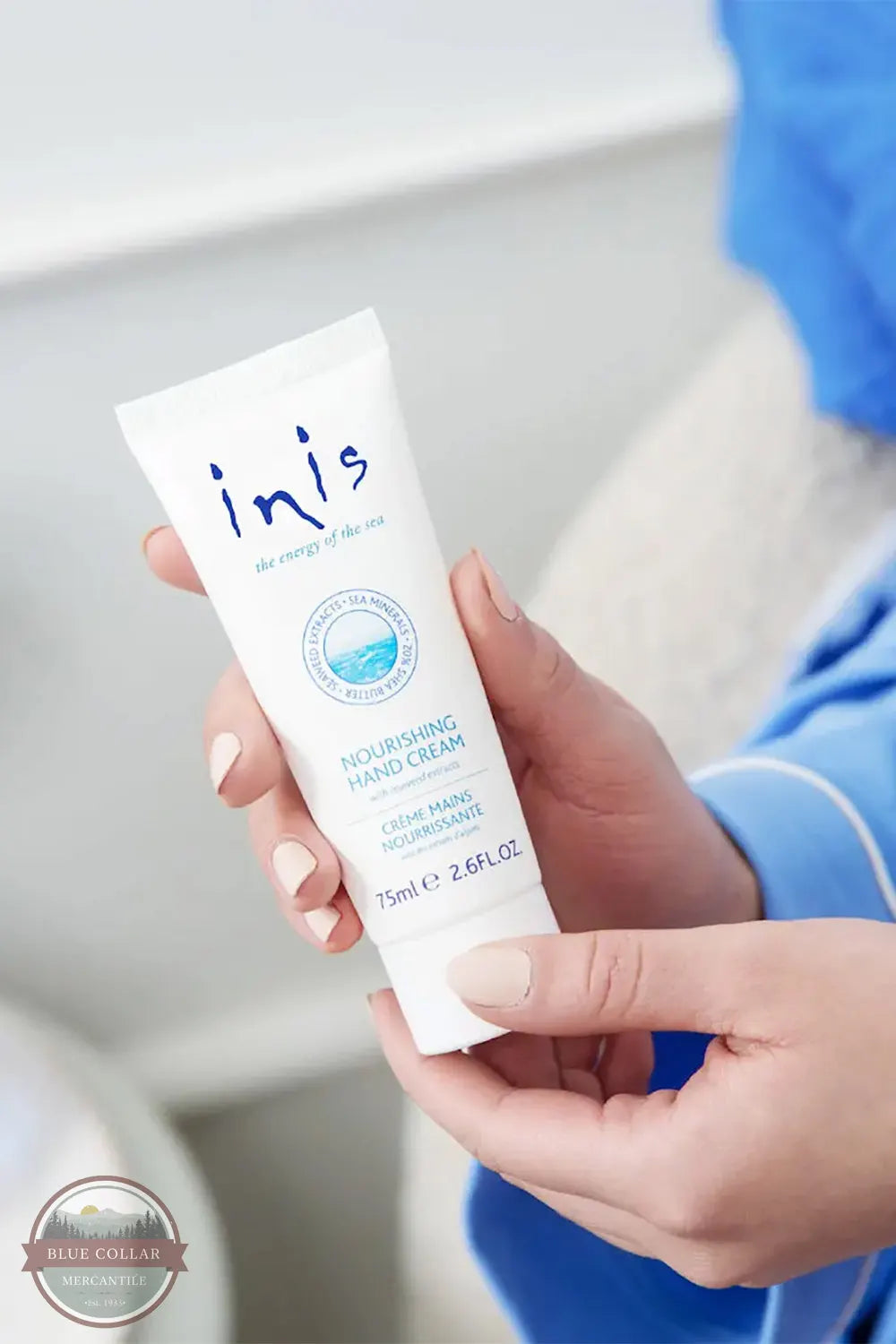 Inis 8015556 Nourishing Hand Cream 2.6FL. OZ. A person holding a white tube of inis Nourishing Hand Cream, with their hands and a blue pajama sleeve visible against a soft, blurred bedroom background.