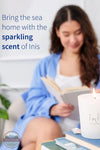 Inis 8017185 Scented Candle 6.7 oz., A woman in a blue robe reads a book in a cozy indoor setting, with a lit white candle in the foreground and text on the left reading 'Bring the sea home with the sparkling scent of Inis'.