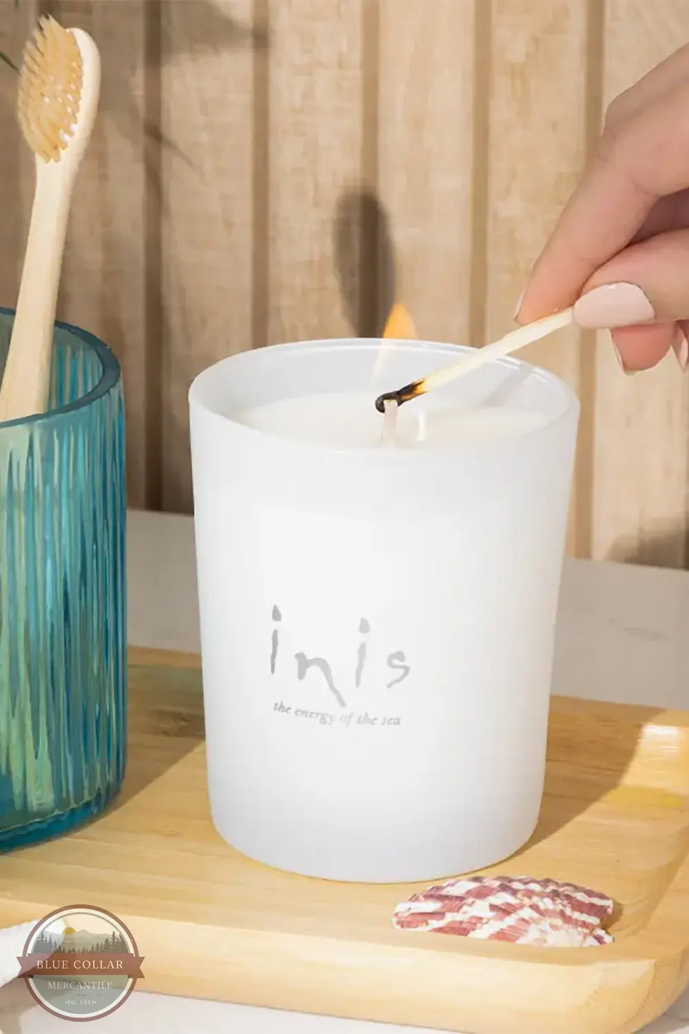 Inis 8017185 Scented Candle 6.7 oz., A frosted white candle labeled 'inis' is lit with a wooden match in a bathroom-like setting, with a hand visible, a blue glass cup with a toothbrush, and a small soap bar on a wooden tray.
