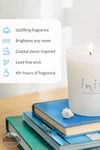 Inis 8017185 Scented Candle 6.7 oz., A white scented candle with a lit flame sits on top of stacked blue books, with a small white flower beside it. A text panel on the left lists product benefits: uplifting fragrance, brightens any room, coastal decor inspired, lead free wick, and 40+ hours of fragrance.