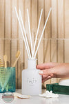Inis 8017222 Fragrance Reed Diffuser 3.3 fl. oz., A frosted white reed diffuser bottle with several white reed straws, held by a hand. The bottle has the brand name 'inis home' printed on it.