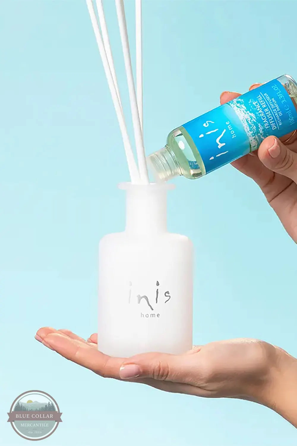 Inis 8017222 Fragrance Reed Diffuser 3.3 fl. oz., A hand pours liquid from a blue bottle into a frosted white reed diffuser bottle, with white reeds extending upward, against a light blue background. The brand name 'inis home' is visible on both the bottle and the diffuser.
