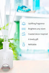 Inis 8017222 Fragrance Reed Diffuser 3.3 fl. oz., A white reed diffuser bottle with white reeds on a light surface, with a blurred bright room in the background and a white info card listing benefits: 'Uplifting fragrance,' 'Brightens any room,' 'Coastal decor inspired,' 'A lovely gift,' and 'Refillable.'.