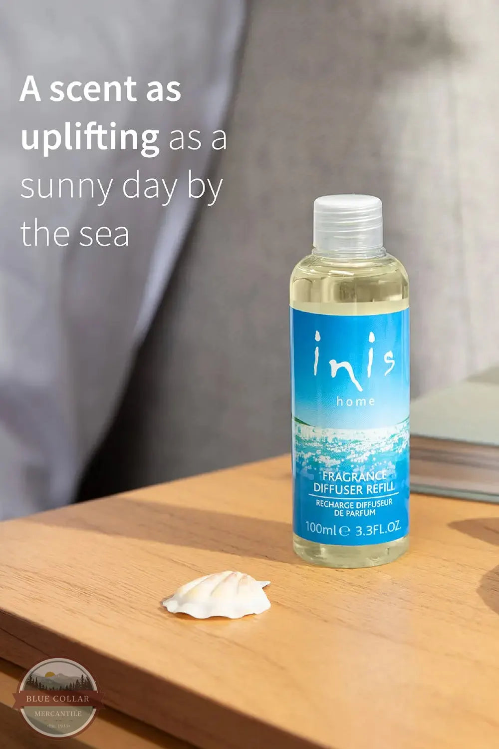 Inis 8017246 Fragrance Diffuser Refill 3.3 fl. oz., A 100ml bottle of Inis Home Escapade Maldives diffuser refill, filled with a pale yellow liquid and featuring a blue tropical wave label, set on a wooden table next to a small white seashell. The background shows a soft-focus gray fabric, with promotional text on the left.