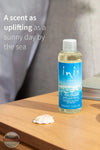 Inis 8017246 Fragrance Diffuser Refill 3.3 fl. oz., A 100ml bottle of Inis Home Escapade Maldives diffuser refill, filled with a pale yellow liquid and featuring a blue tropical wave label, set on a wooden table next to a small white seashell. The background shows a soft-focus gray fabric, with promotional text on the left.
