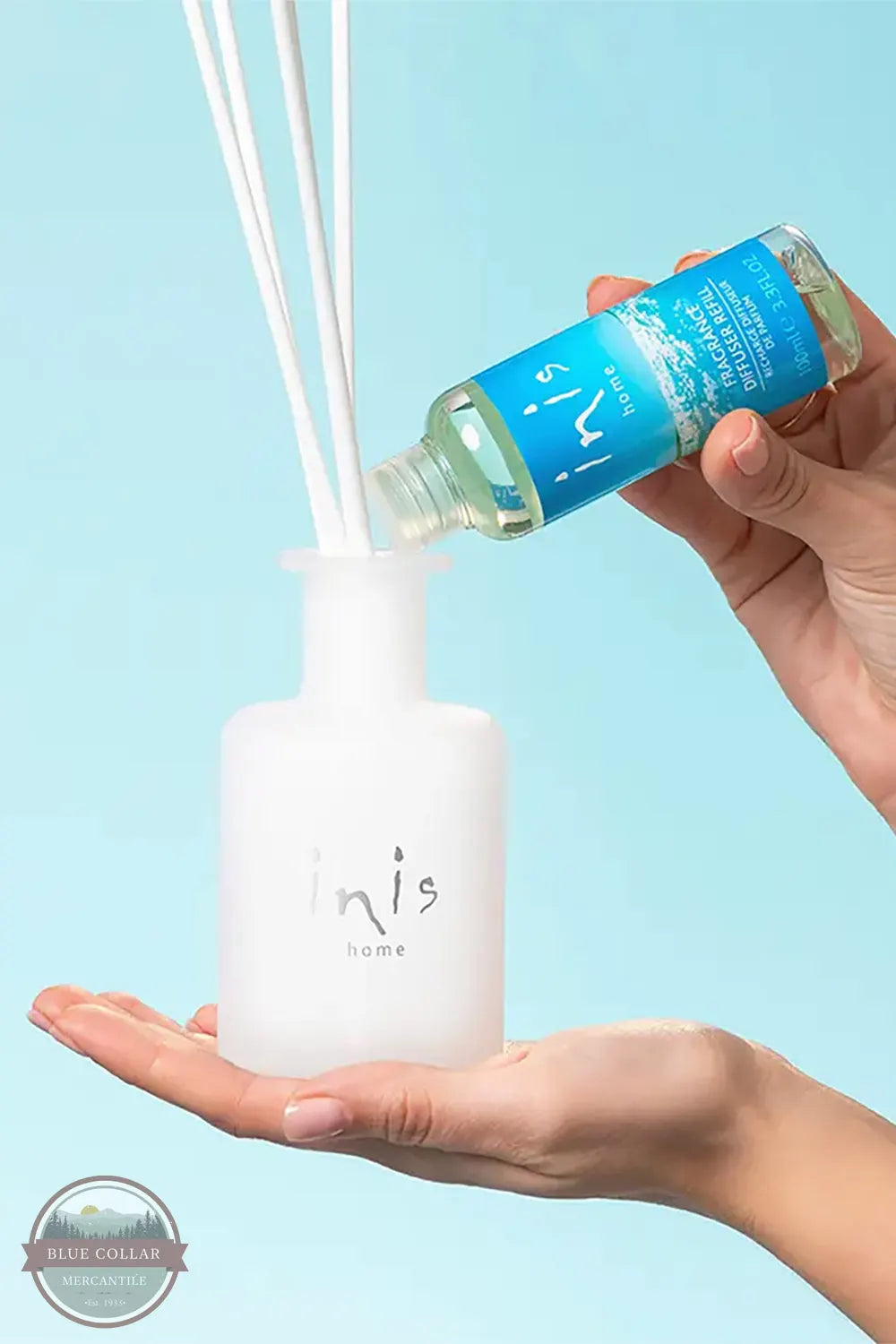 Inis 8017246 Fragrance Diffuser Refill 3.3 fl. oz., A hand pours blue liquid from a small bottle into a frosted white reed diffuser bottle held in another hand, with white reeds extending upward against a light blue background. The bottle label shows the brand name Inis Home.