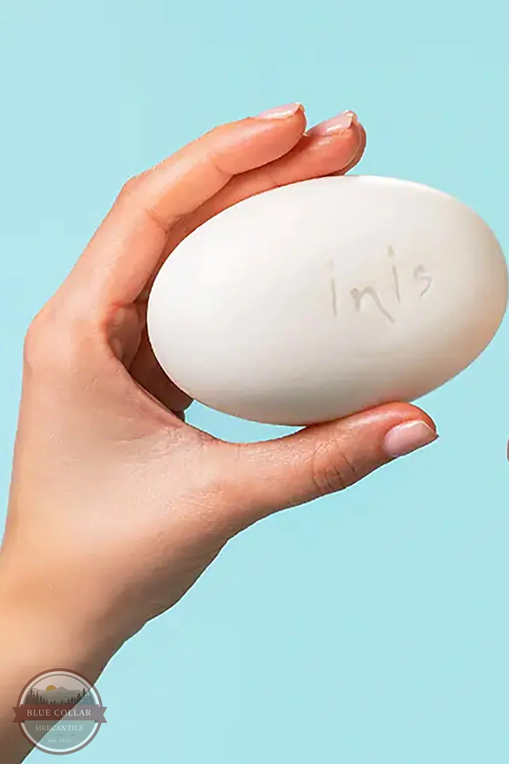 Inis 8018618 Sea Mineral Soap Bar, Large A hand holding a large white oval bar of soap with the brand name 'inis' written on it, set against a solid light blue background.