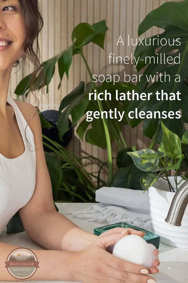 Inis 8018618 Sea Mineral Soap Bar, Large A woman in a white ribbed tank top holds a white bar of soap in her hands at a sink, with large green plants and a faucet in the background. White text on the right reads, 'A luxurious finely-milled soap bar with a rich lather that gently cleanses.