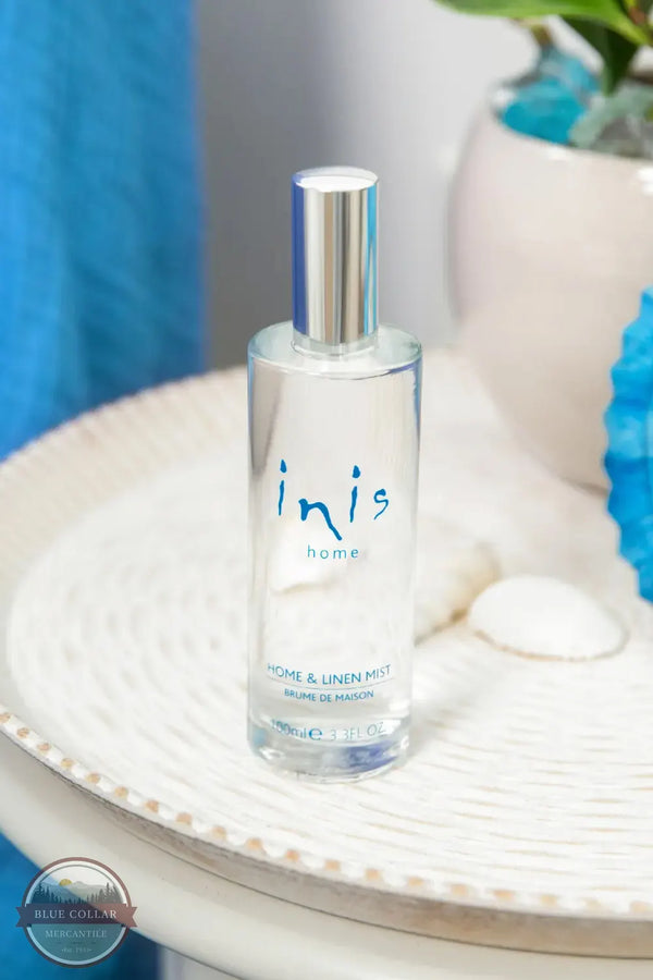 Inis 8020901 Home &amp; Linen Mist 3.3fl. oz., A clear glass bottle of Inis Home Home & Linen Mist with a silver cap, displayed on a white textured tray with seashells and blue decorative elements in the background.