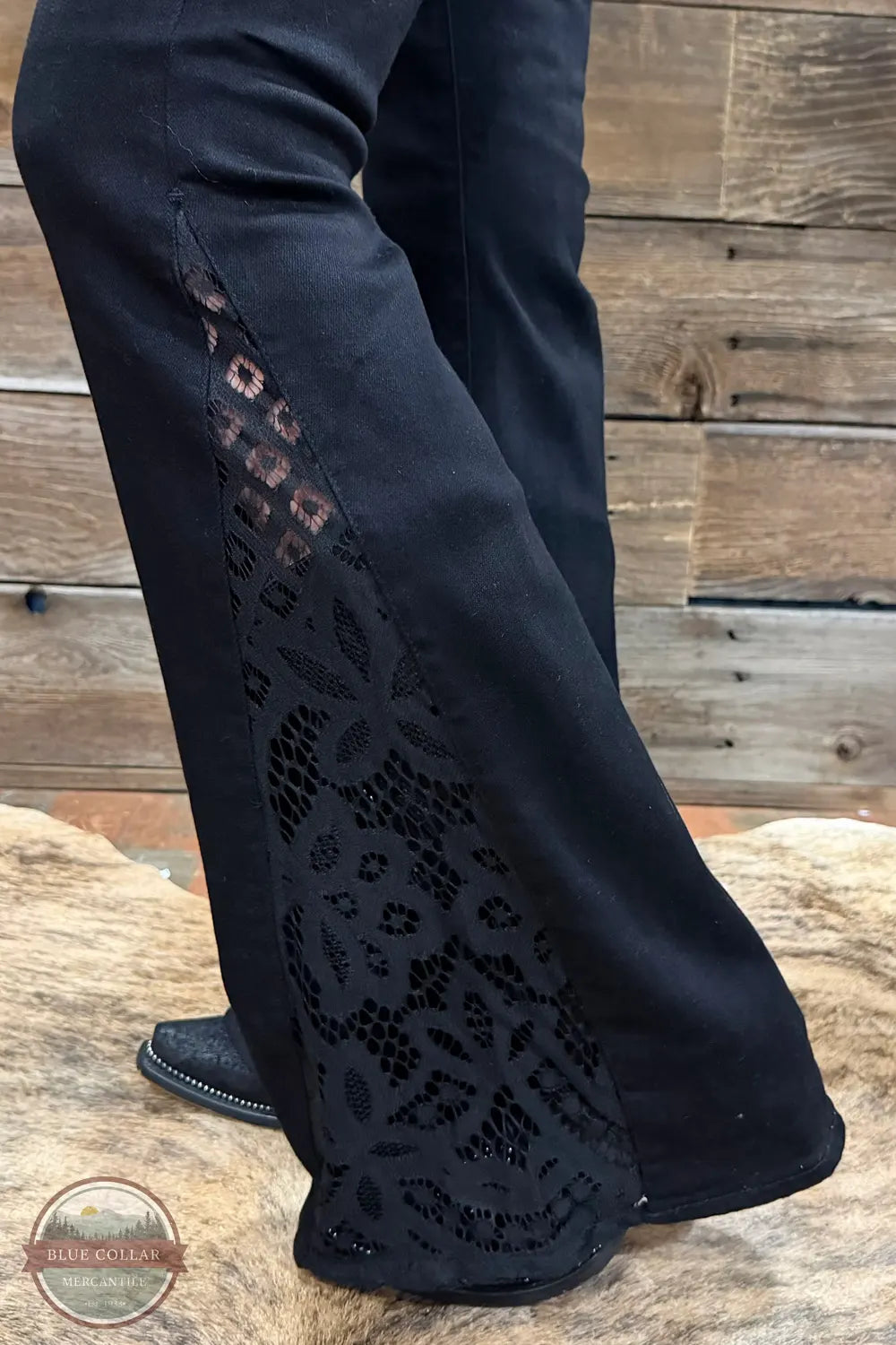 Judy Blue 881177REG High Waist Black Jaqueline Flare Jean With Lace, Black wide-leg pants with floral lace applique panels on the outer sides, shown on a model standing on a rustic wooden surface.
