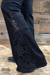 Judy Blue 881177REG High Waist Black Jaqueline Flare Jean With Lace, Black wide-leg pants with floral lace applique panels on the outer sides, shown on a model standing on a rustic wooden surface.