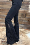 Judy Blue 881177REG High Waist Black Jaqueline Flare Jean With Lace, Black flare pants with a black lace overlay detail on the lower leg, shown on a model standing in front of a rustic wooden wall and fur rug.